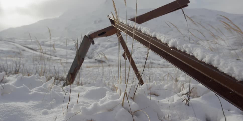 A rusty frame sticks out of the snow as wind blows the grass