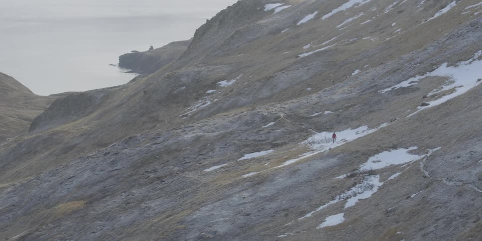 A hiker walks up a mountainside with patches of snow
