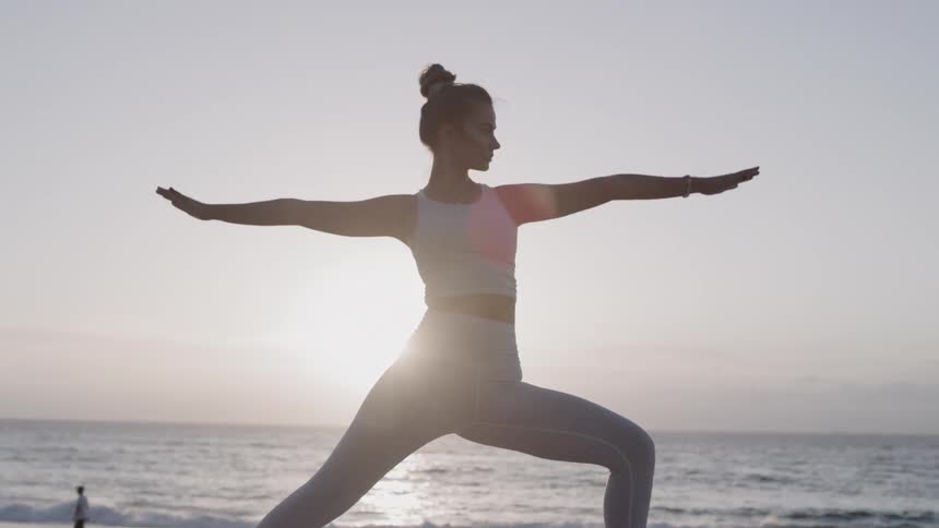 A woman does yoga on a mat on the beach at sunset
