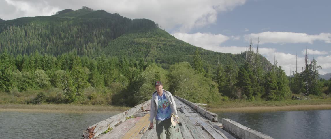 A man carrying a skateboard and walking over a wooden bridge that spans a body of water in the mountains
