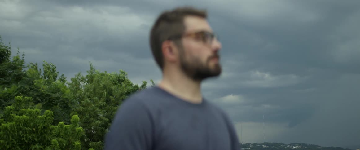 A man with glasses and a beard is standing outside looking at something in the distance