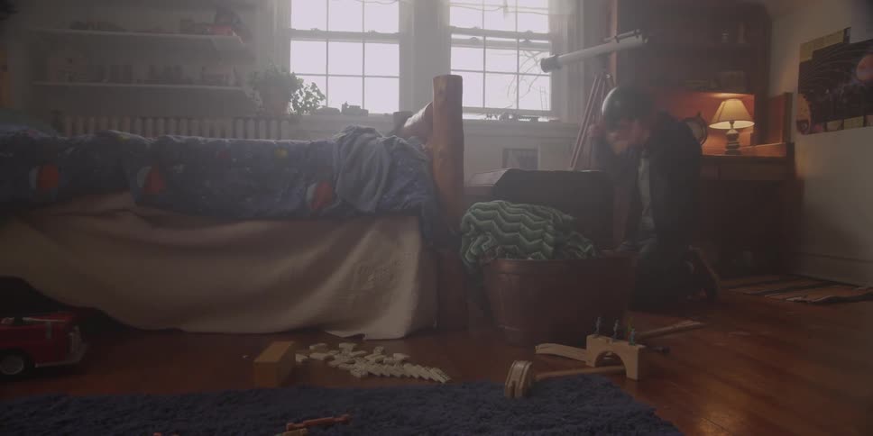 A boy wearing a helmet kneeling down in front of a chest in a bedroom,  taking off the helmet and opening the chest