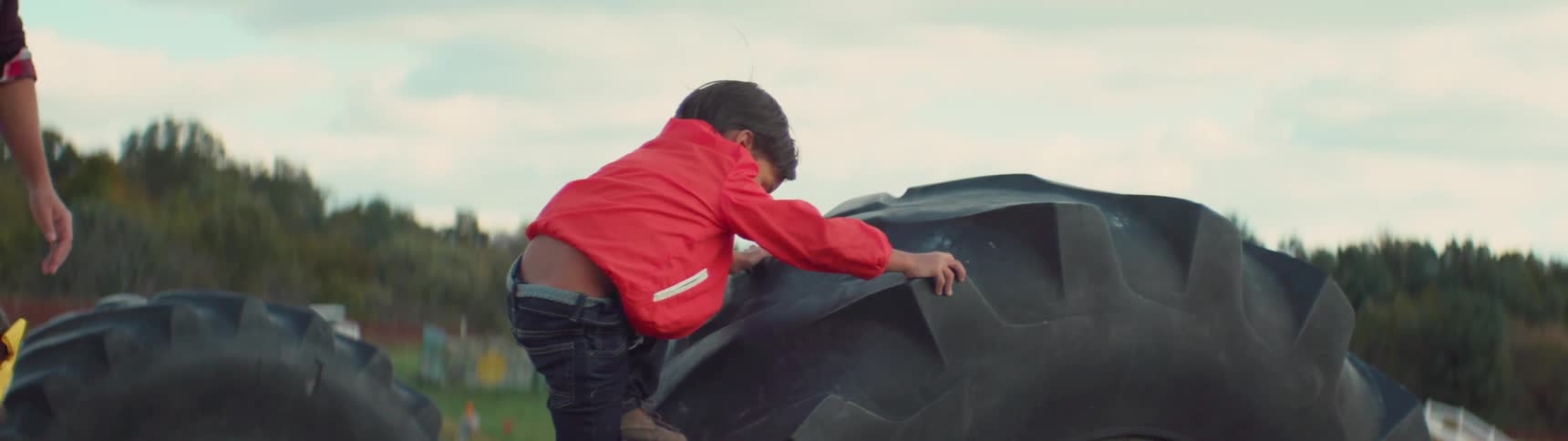 A child is climbing on top of a large tractor tire as his father helps him