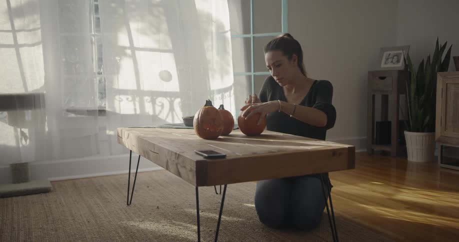 A woman kneels at a coffee table and carves pumpkins
