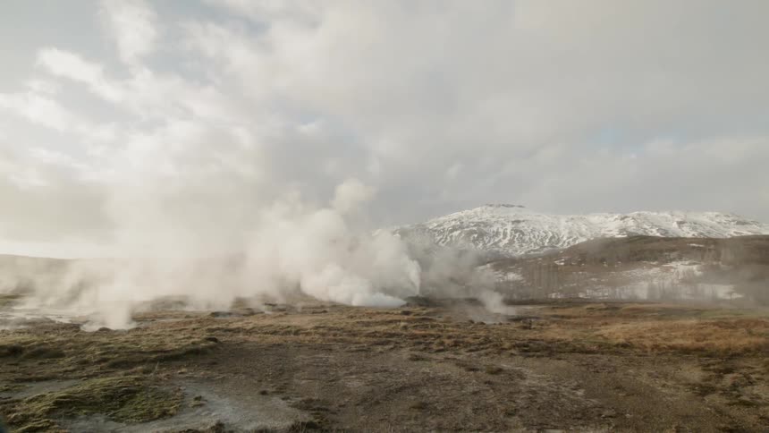 A hot spring is releasing steam in a wide open plain with a snow covered hill in the background