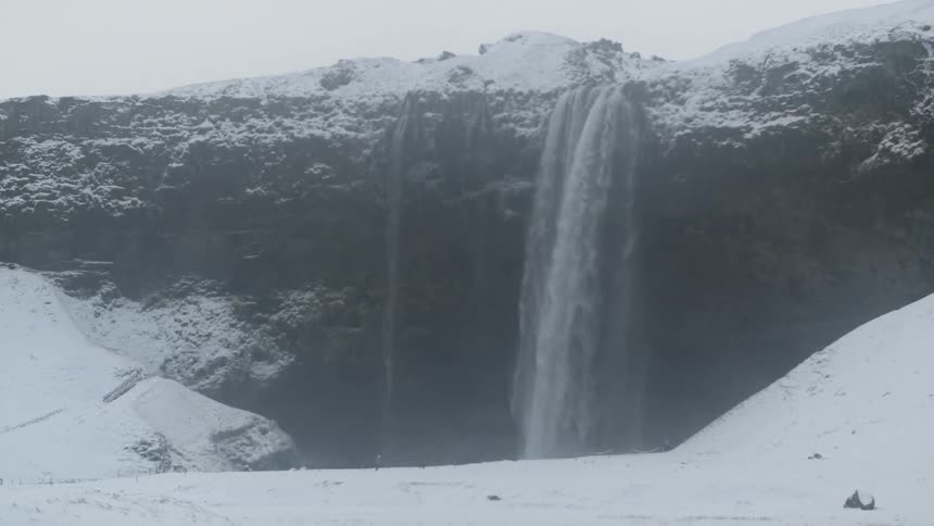 A large waterfall is falling down a snow covered cliff