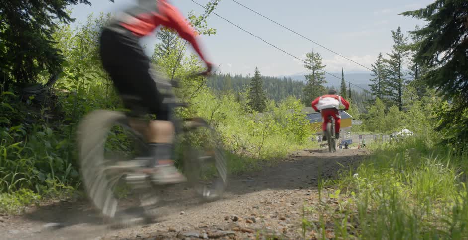 Two people ride bikes on a dirt path surrounded by trees and bushes