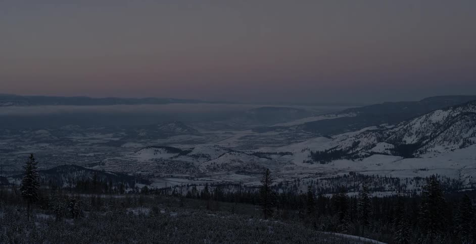 Dusk is settling over a valley and snow-covered mountains in winter