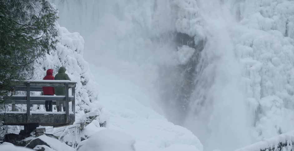 Water and snow fall off a tall cliff as two people look on
