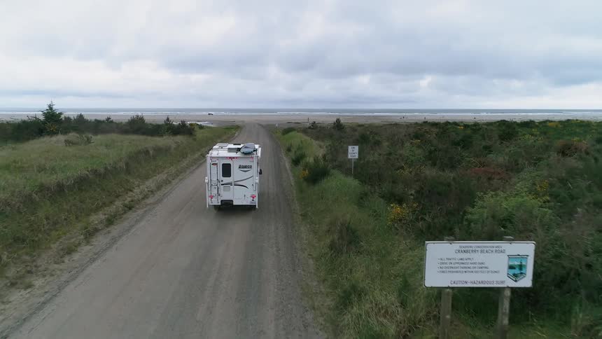 A camper trailer is being driven on a dirt road in a coastal setting on a cloudy day