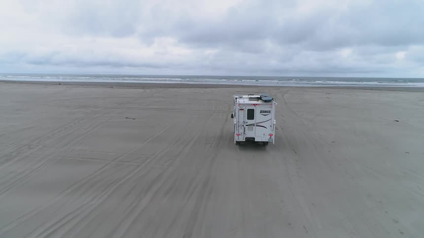 A camper trailer is being driven on the beach toward the ocean on a cloudy day