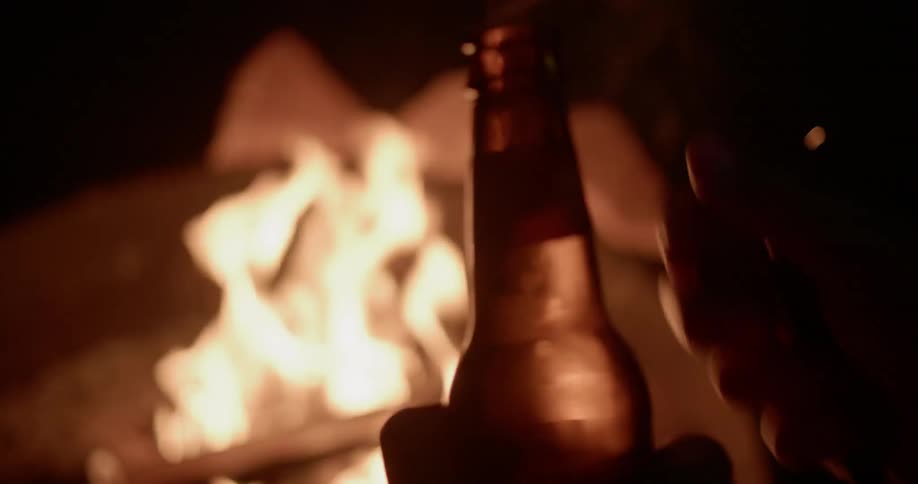 A man is opening a beer bottle as he sits by a campfire at night