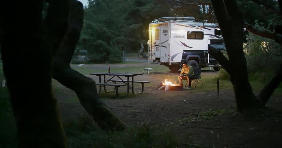 A man sits at a campfire with his RV parked behind him