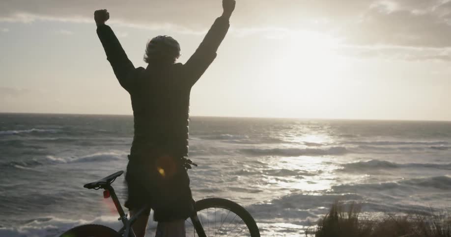 A bicyclist raises his arms in celebration on a beach at sunset