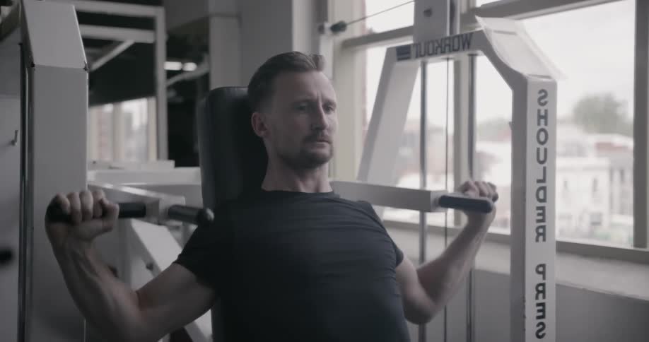 A man is working out on a shoulder press machine in a gym during the day
