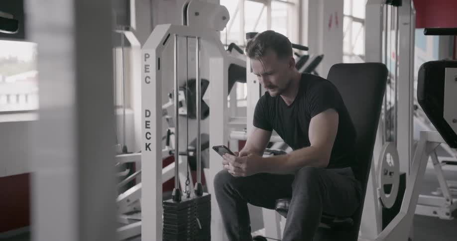 A man types on his phone while sitting in a gym