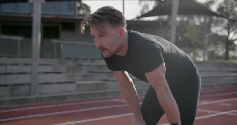 A man is resting his hands on his knees and breathing heavily outside on a track during the day