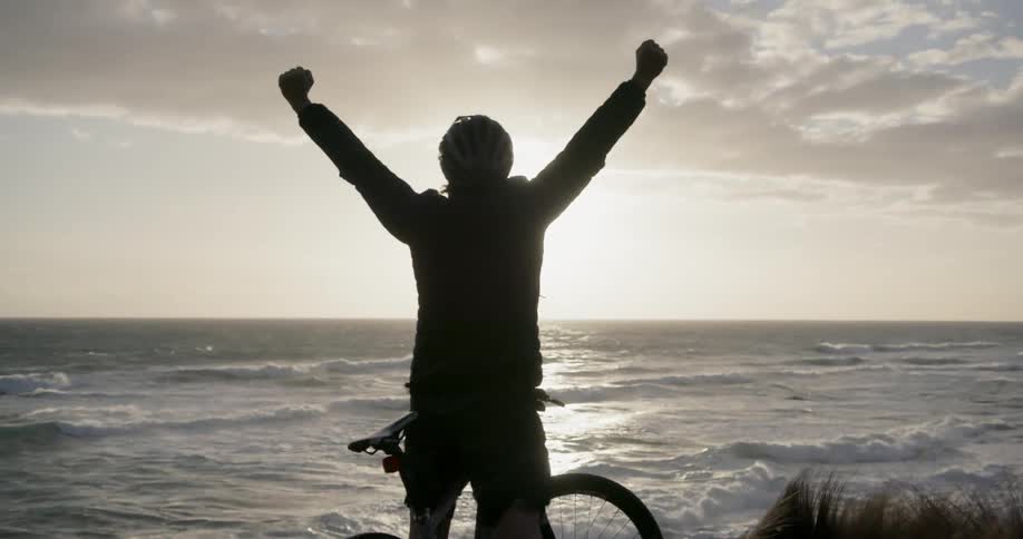 A bicyclist raises his arms in celebration on a beach at sunset