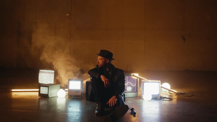 A man in a black hat sits in front of vintage tvs and computers that are flickering static