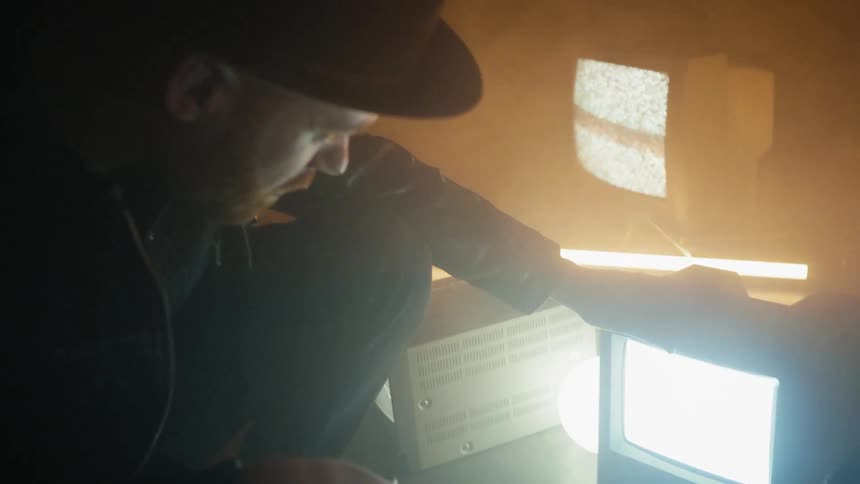 A man crouches next to tvs on the floor of a smoky room