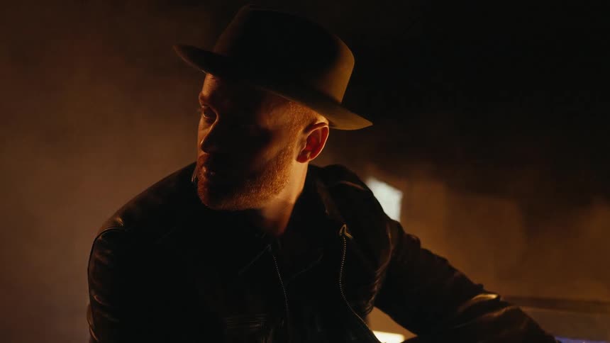 A man sits in a dim and smoky room with flickering television screens