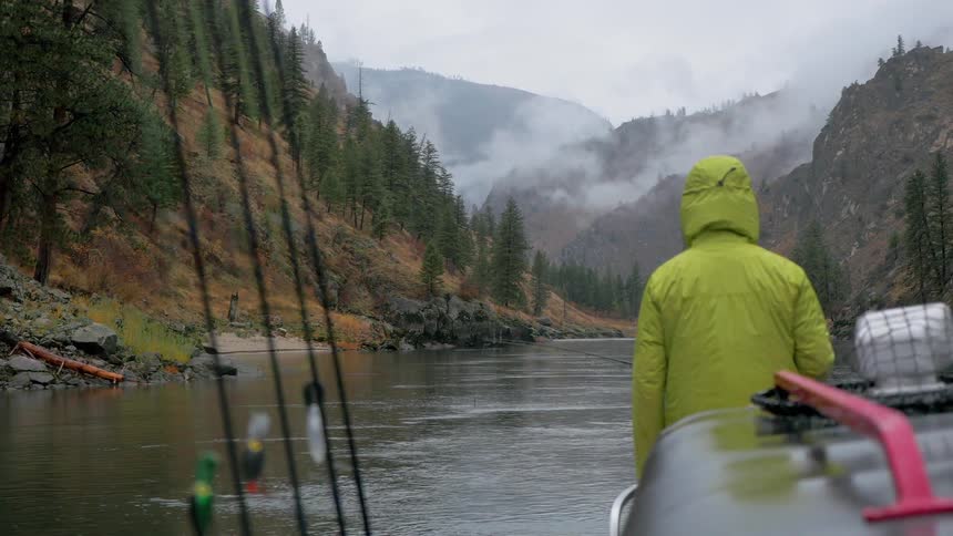 A fisherman stands at the front of a boat traveling down a river