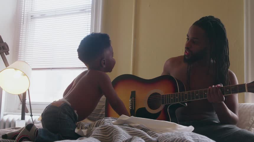 A man and young boy play with a guitar on a bed