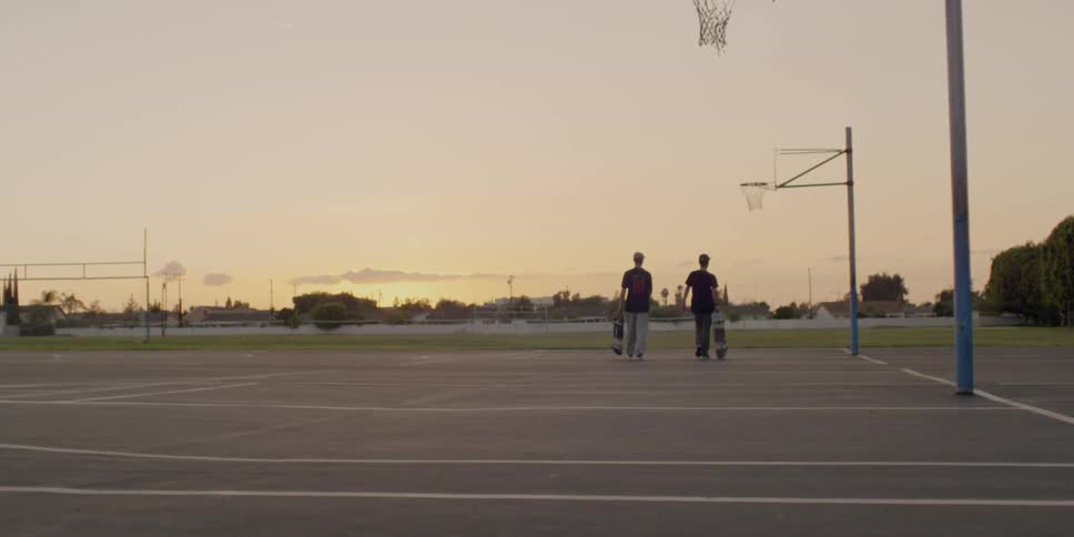 Two boys carrying skateboards walk on a basketball court at sunset