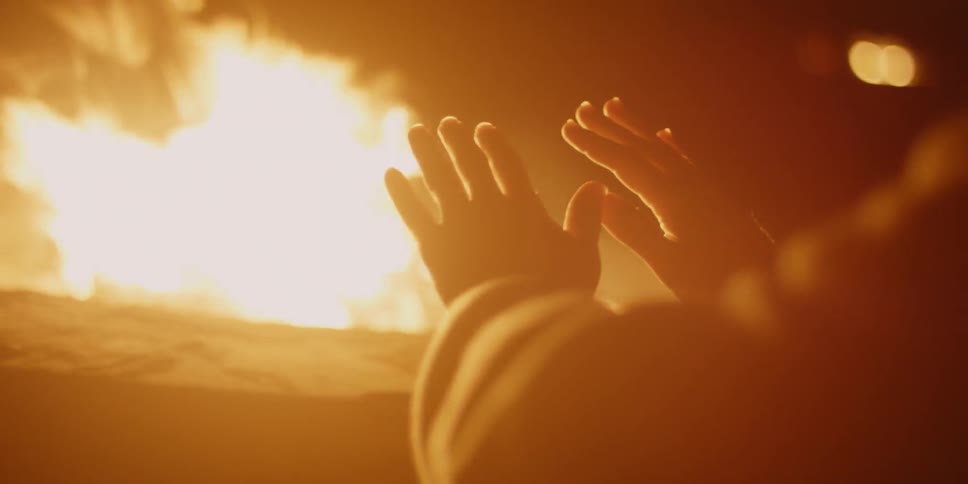A man is rubbing his hands as he sits near a campfire at night