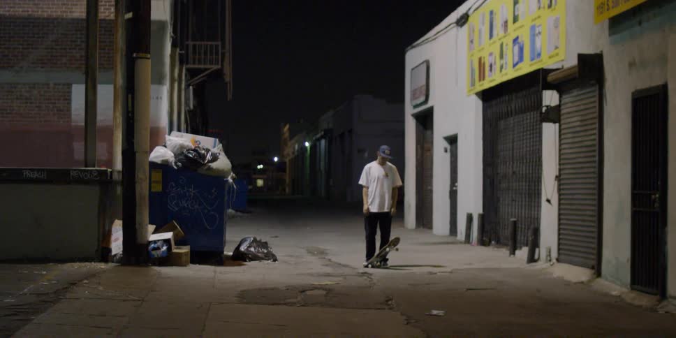 A man puts his skateboard on the ground in an alley near a dumpster