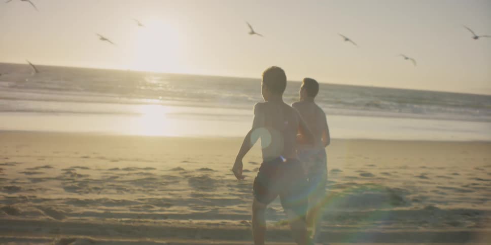 Two teenagers are running on a beach towards the waves at sunset