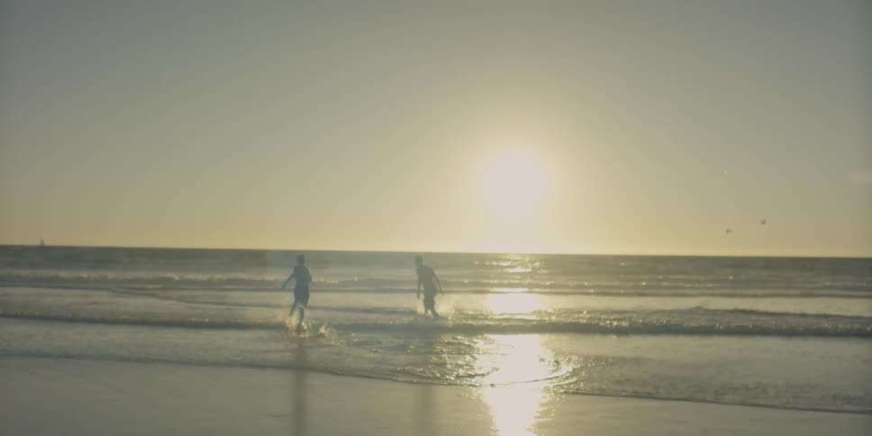 Two boys are playing as they run into the surf on a beach at sunset