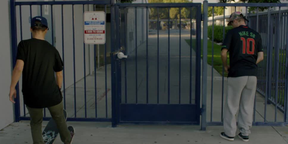 Teenagers on skateboards are approaching a gated fence in a neighborhood during daylight