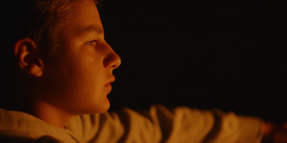 A boy is talking as he sits near a campfire at night