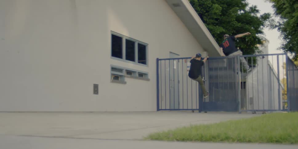 Two people climb over a fence with skateboards