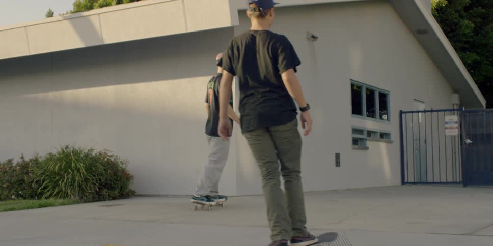 Two men ride their skateboards towards the fence in the lot