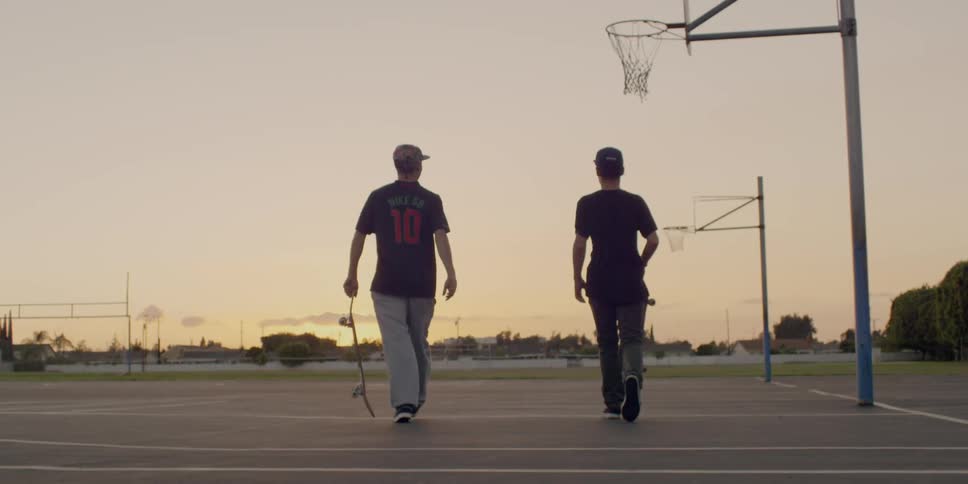Two teenagers are carrying skateboards across a basketball court outdoors at dusk