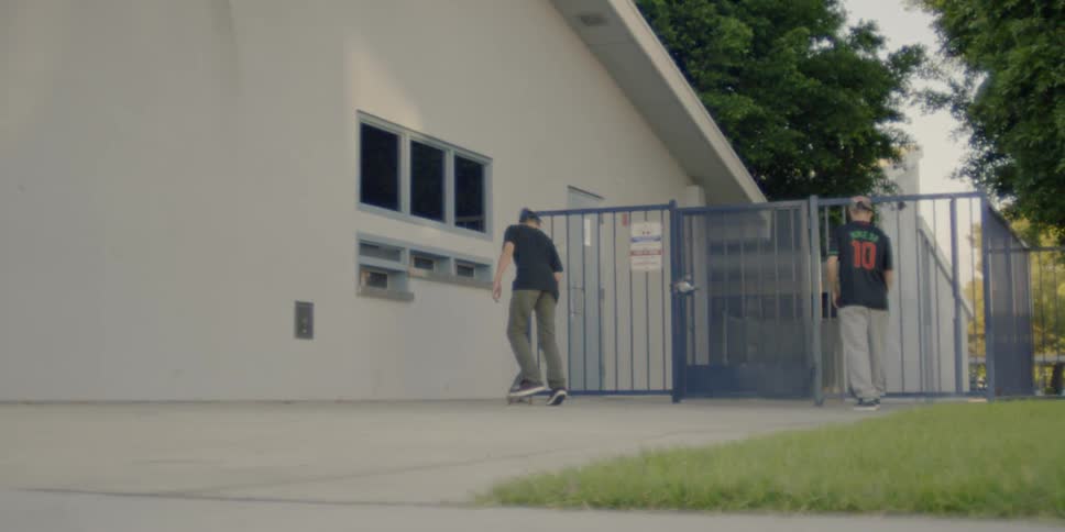Two men ride up to a fence with their skateboards