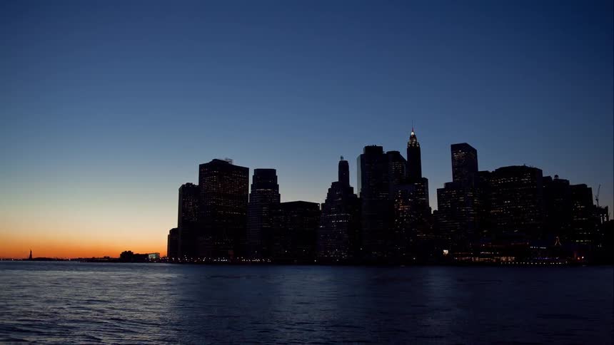 Sun sets behind a flowing body of water and buildings light up at night