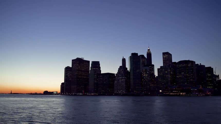 Sun sets behind a flowing body of water and buildings light up at night