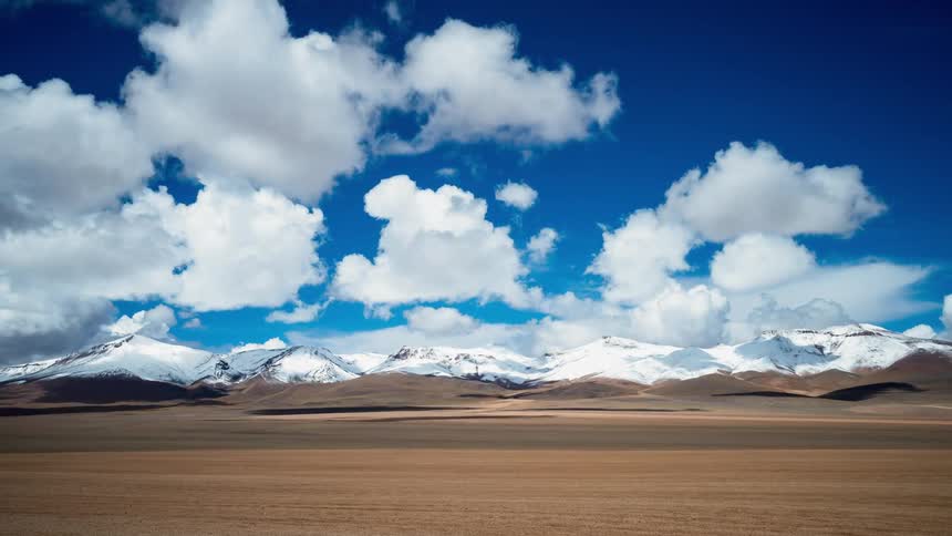 White clouds plume and pass over an open field near a snow covered mountain