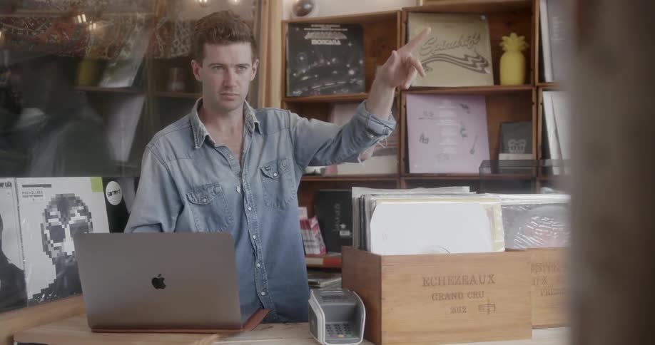 A man in a record store is gesturing and  working at a computer