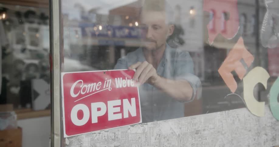 Man turning around a sign to indicate a shop is open as traffic passes by