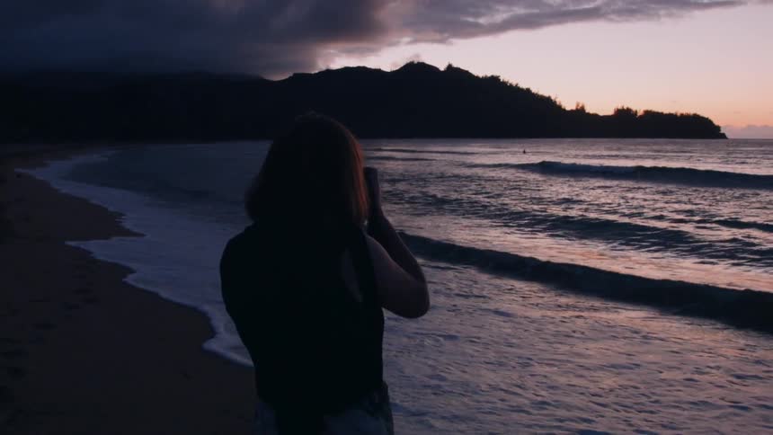 Woman taking pictures of the sea coming ashore on a cloudy sunset