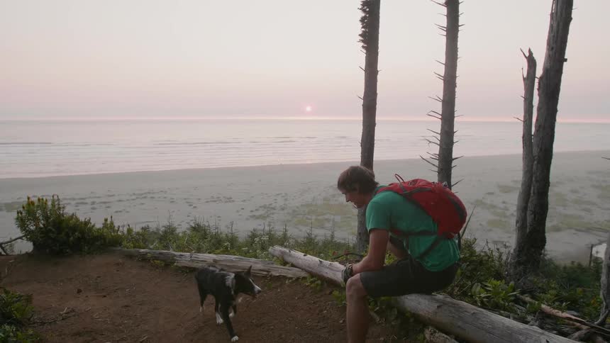 A man holding a helmet plays fetch with his dog near the beach