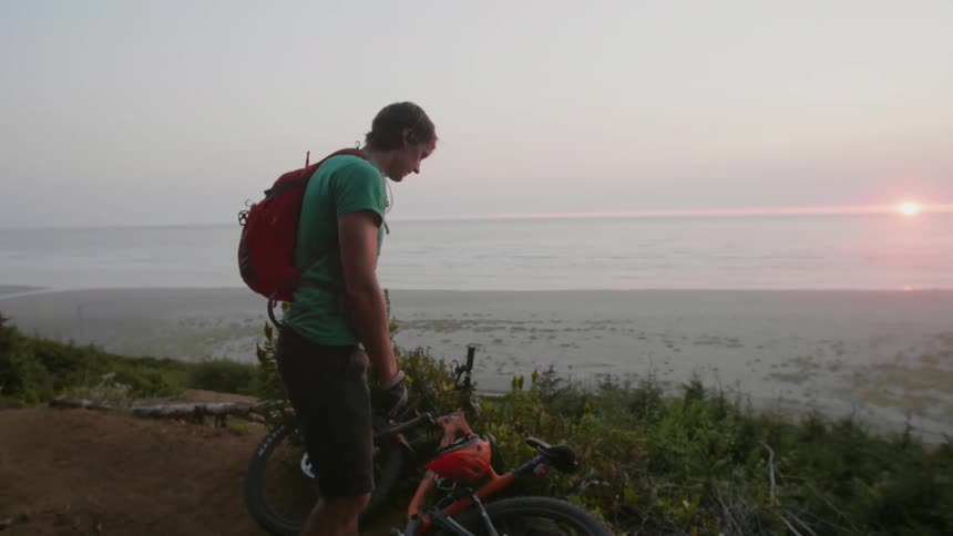 A bicyclist stands atop a hill with a dog and looks out towards the water