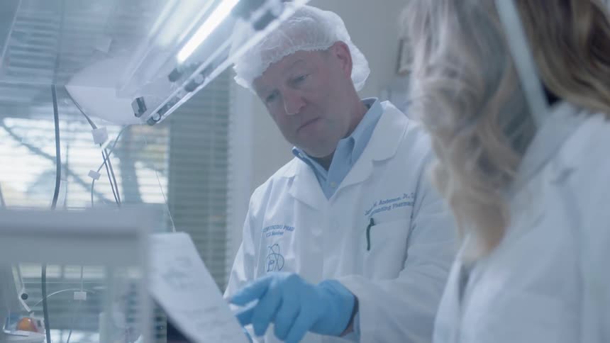 Man and woman wearing lab coat and gloves look at a piece of paper while standing in a lab