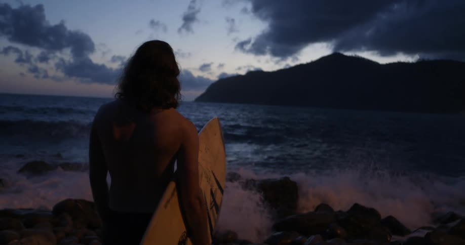 A man stands with his surfboard watching the sunset