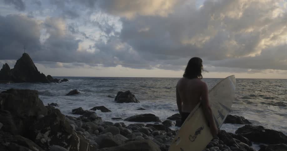 A surfer stands on a rocky beach at sunset