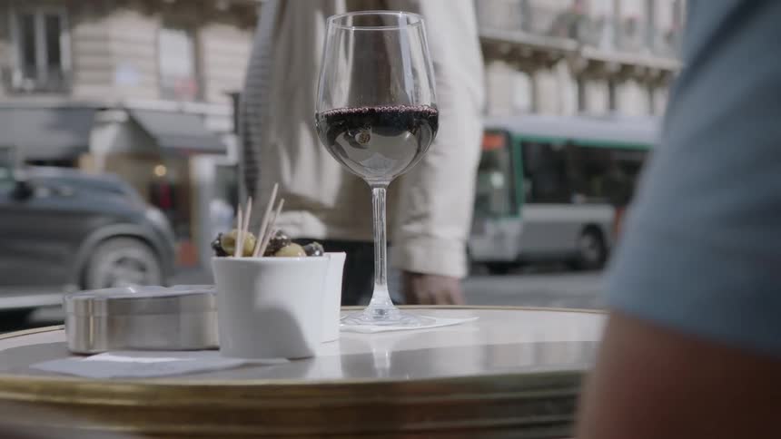 Person sits at a table with a glass of wine on it outside with people and vehicles passing by